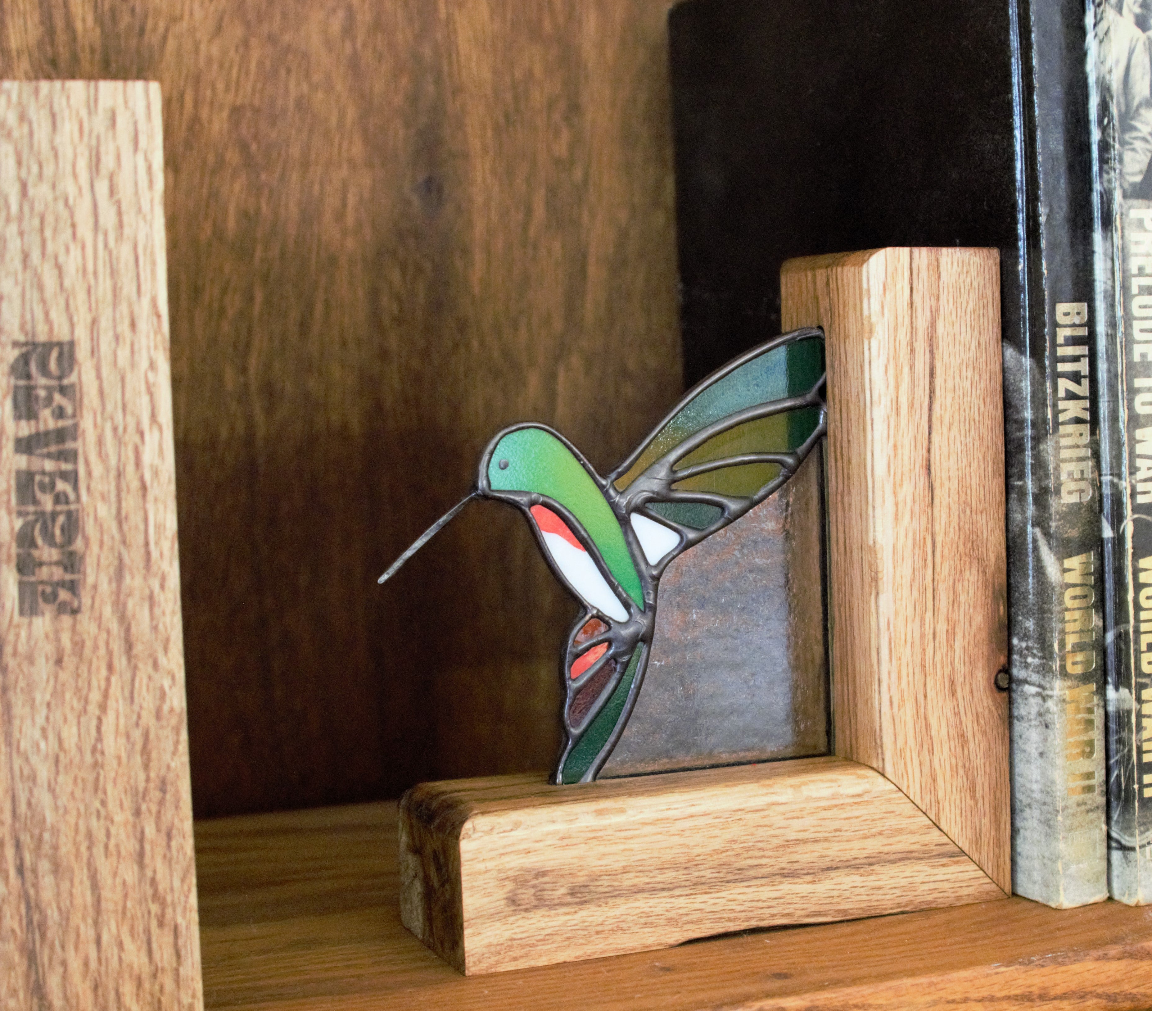 Handmade stained glass hummingbird bookend in jewel-toned blue and green, on a wood shelf
